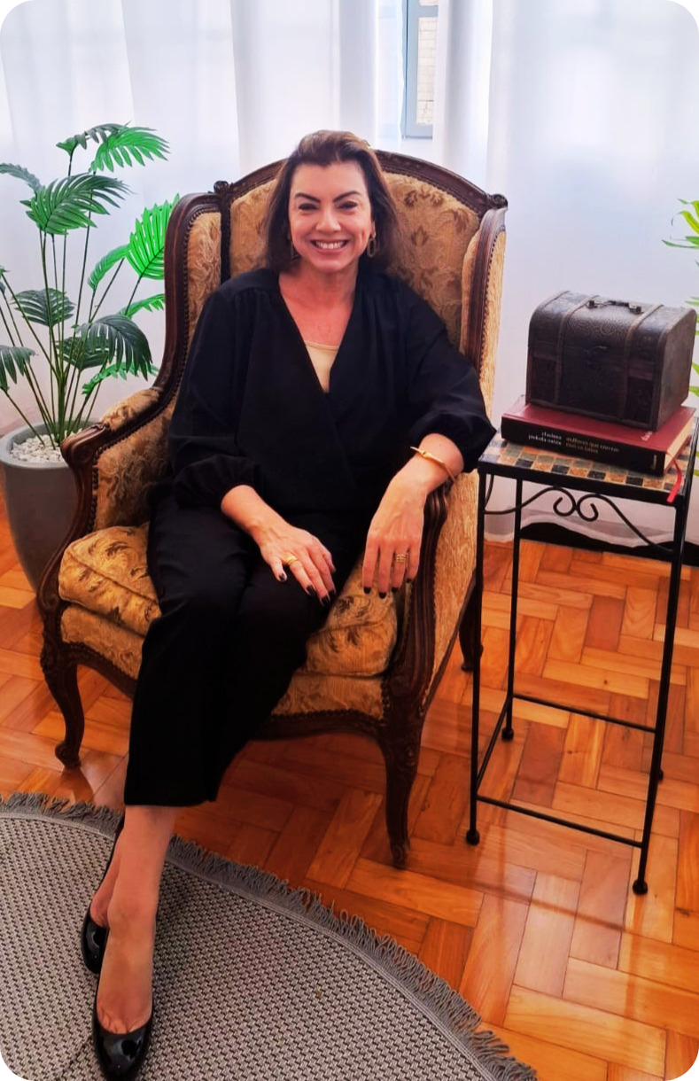 Mulher sorridente de terno preto e camisa branca, cabelo castanho, fundo claro, usando brincos discretos.